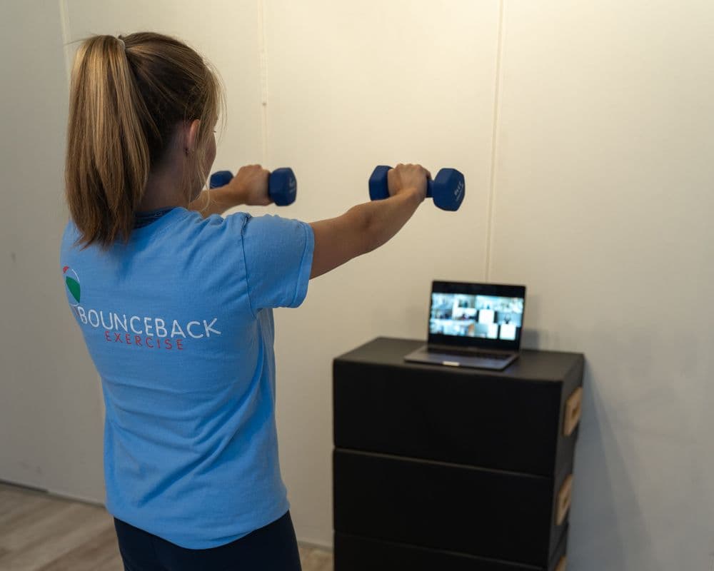 A woman exercising indoors using weights while a virtual class can be seen on her laptop.