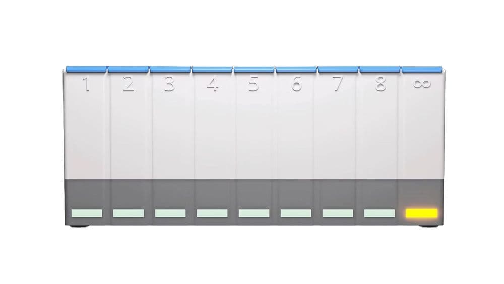 A photo of the white, grey and blue MedpeR dosette box. There is a yellowish orange light on the slot in the bottom right corner. 
