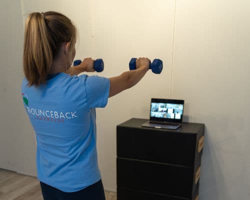 A woman exercising indoors using weights while a virtual class can be seen on her laptop.