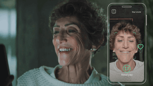An older woman smiling at her phone. Her phone is shown next to her and she is smiling on the screen. There us a smiley face below her and the words 'Good job!'.