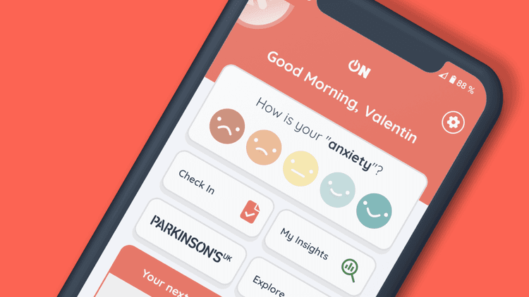 A close-up photo of a phone with the Parkinson's On app on the screen. You can see the homepage, including the words "Good Morning, Valentin", an anxiety rating, a check-in button and links to your Insights and Explore pages.