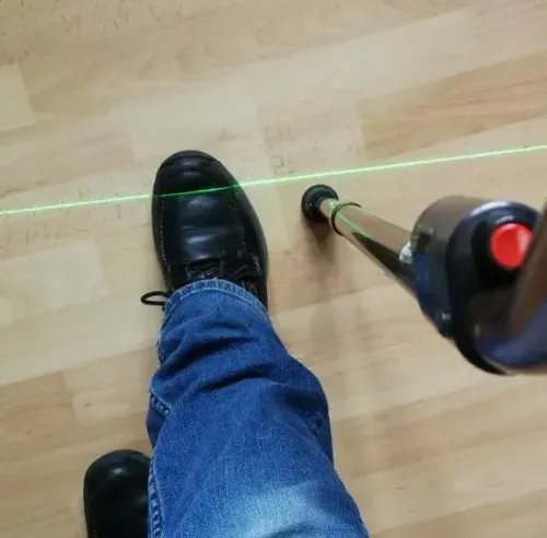A photo of a green laser projected on the floor overlapping a man's foot as he holds the LaserCane.